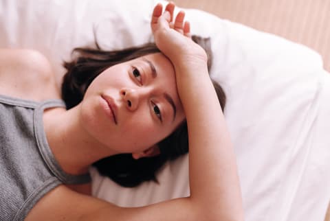 Brunette woman lying on her bed wearing a grey tank top, looking tired or distressed, resting her forearm on her forehead.